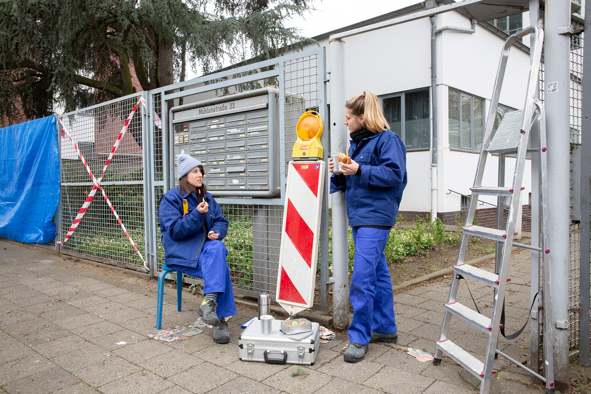 Zwei Frauen in blauen Arbeitsanzügen machen vor einem Zaun Pause